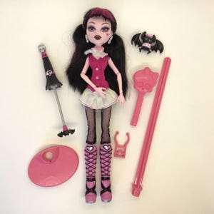 Peut inclure: Poupée avec des cheveux noirs et des mèches roses, vêtue d'une tenue rose et blanche, de bas résille et de bottes roses. Accessoires : une chauve-souris, un objet en forme de crâne, un socle rose et un parapluie noir avec un motif de chauve-souris. Poupée de collection.