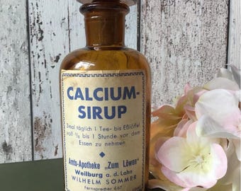 May include: An antique amber glass bottle with a black cap and a label that reads "CALCIUM-SIRUP." The label also includes German text. The bottle is likely from a pharmacy, with the name "Amts-Apotheke Zum Löwen" visible.