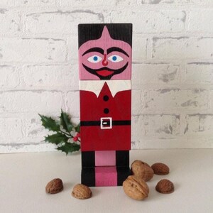 Puede incluir: Una figura navideña de madera pintada en rojo, negro, rosa y blanco, que se asemeja a un personaje tipo Papá Noel. La figura está hecha de bloques apilados y tiene una cara con ojos azules y un bigote. Nueces y ramitas de acebo en primer plano.