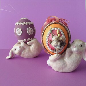 May include: Two ceramic bunny figurines holding decorative Easter eggs. One egg is purple with white floral details, the other is orange with a pink floral design and a pink ribbon. The bunnies are white.