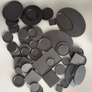 Pode incluir: Uma variedade de bandejas e pratos de metal preto em várias formas e tamanhos. A coleção inclui peças redondas, ovais, quadradas e retangulares, algumas com bordas elevadas ou centros rebaixados. As superfícies têm um acabamento fosco.