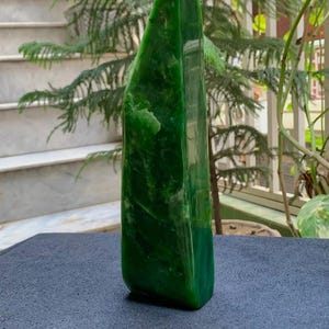 May include: A polished, vibrant green jade sculpture with a smooth, glossy finish. The stone has a unique, asymmetrical shape and stands upright on a dark surface. The sculpture's color varies from light to dark green.