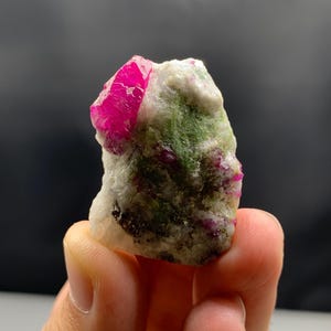 Op de afbeelding: Een natuurlijk mineraal specimen met een levendig, felroze robijnkristal. De robijn is gezet in een matrix van witte en groene mineralen, wat een opvallend contrast creëert. De ruwe, ongepolijste textuur benadrukt de ruwe schoonheid van de edelsteen.