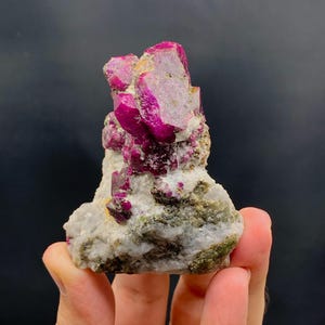 Puede incluir: Un grupo de vibrantes cristales de rubí de color rosa intenso sobre una matriz de piedra blanca y verde. Los cristales de rubí tienen una textura rugosa y natural, y la matriz tiene un aspecto moteado. Una llamativa exhibición de belleza mineral natural.