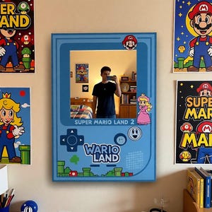 May include: A blue mirror in the shape of a Super Mario Land 2 Game Boy. The mirror has a rectangular shape with rounded corners and features Mario and Princess Peach stickers. The words "Super Mario Land 2" and "Wario Land" are printed on the mirror.
