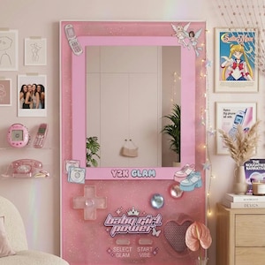 Op de afbeelding: Een roze Y2K-thema spiegel met een retro videogame controller ontwerp. De spiegel heeft de tekst "Y2K GLAM" en "baby girl power". Decoratieve elementen zijn onder meer een telefoon, engelstickers en een hartvormige luidspreker.
