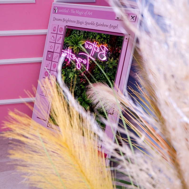 K&ouml;nnte beinhalten: Ein rosa gerahmtes Display mit dem Text "untitled - The Magic of Art" und "Dreams Brightness Magic Sparkle Rainbow Angel". Im Inneren des Rahmens befindet sich ein Neonschild mit der Aufschrift "Better". Der Hintergrund ist eine Wand aus Pflanzen. Pampasgras befindet sich im Vordergrund.