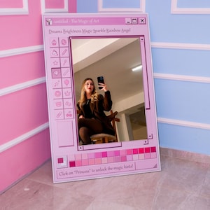 K&ouml;nnte beinhalten: Ein rosa gerahmter Spiegel mit einem Design einer Computer-Oberfl&auml;che. Der Spiegel reflektiert eine Person, die ein Telefon h&auml;lt. Der Text auf dem Rahmen lautet "untitled - The Magic of Art" und "Dreams Brightness Magic Sparkle Rainbow Angel".