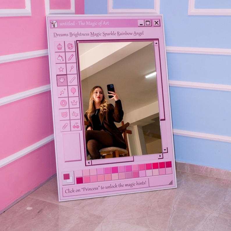 K&ouml;nnte beinhalten: Ein rosa gerahmter Spiegel mit einem Design einer Computer-Oberfl&auml;che. Der Spiegel reflektiert eine Person, die ein Telefon h&auml;lt. Der Text auf dem Rahmen lautet "untitled - The Magic of Art" und "Dreams Brightness Magic Sparkle Rainbow Angel".