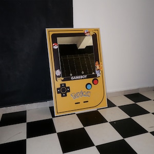Puede incluir: Un espejo enmarcado en amarillo dise&ntilde;ado como una Gameboy, con la palabra "GAMEBOY" y el logo de "Pok&eacute;mon". El espejo tiene un borde negro y est&aacute; decorado con personajes de Pok&eacute;mon y una Pok&eacute;ball.