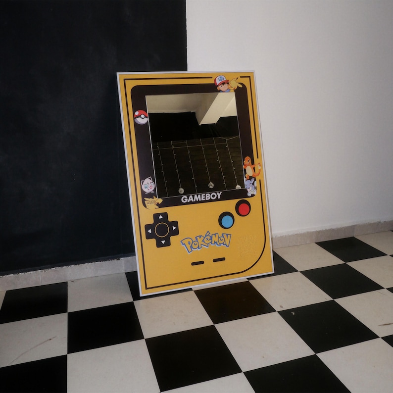 Puede incluir: Un espejo enmarcado en amarillo dise&ntilde;ado como una Gameboy, con la palabra "GAMEBOY" y el logo de "Pok&eacute;mon". El espejo tiene un borde negro y est&aacute; decorado con personajes de Pok&eacute;mon y una Pok&eacute;ball.