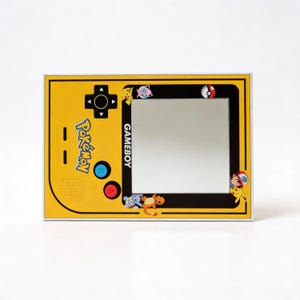 Puede incluir: Un espejo amarillo con tem&aacute;tica de Gameboy con un borde negro. El espejo presenta las palabras "Pok&eacute;mon" y "GAMEBOY" en texto negro. Varios personajes de Pok&eacute;mon se muestran alrededor del borde del espejo, incluyendo Pikachu, Charmander y Squirtle.