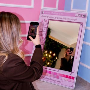 K&ouml;nnte beinhalten: Ein rosa gerahmter Spiegel mit dem Text "untitled - The Magic of Art" und "Dreams Brightness Magic Sparkle Rainbow Angel". Der Spiegel hat ein dekoratives Design und wird mit einem Smartphone fotografiert.