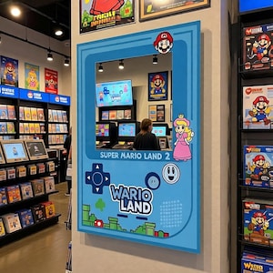 May include: A light blue wall decoration featuring the Super Mario Land 2: Wario Land game. The decoration includes images of Mario, Princess Peach, and game controller symbols. The text "SUPER MARIO LAND 2" and "WARIO LAND" are also visible.