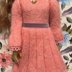 May include: A pink knitted doll dress with a purple velvet belt and a crocheted lace trim. The dress has a round neckline and long sleeves.