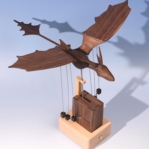 May include: A dark brown wooden dragon automaton with articulated wings and a tail, mounted on a light-colored wooden base. The dragon is connected to a mechanical box by thin metal rods and wires, allowing for movement of the wings and body parts.