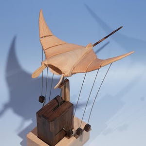 May include: A wooden sculpture of a manta ray with a dark wooden box base. The ray is light brown with a dark brown rod extending from its back. The base is light brown with a dark brown box suspended by wires.