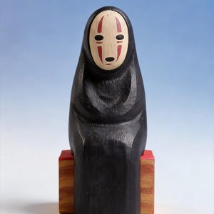 May include: A carved wooden figure of the character No-Face from the animated film Spirited Away. The figure is black with a white face, red markings, and is seated on a red and brown wooden block. The background is a gradient of blue and white.