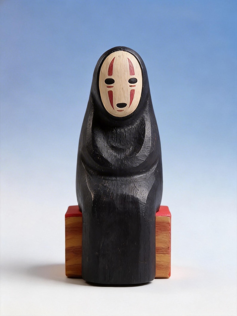 May include: A carved wooden figure of the character No-Face from the animated film Spirited Away. The figure is black with a white face, red markings, and is seated on a red and brown wooden block. The background is a gradient of blue and white.