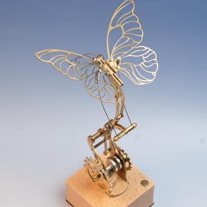 Puede incluir: Escultura de mariposa mecánica en color latón, con intrincados detalles de engranajes y palancas. Las alas de la mariposa están delicadamente cortadas, y la escultura está montada sobre una base de madera. El diseño evoca una estética steampunk.