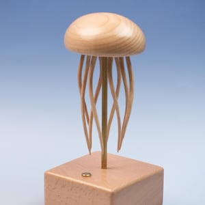 Könnte beinhalten: Holzskulptur einer Qualle mit einem abgerundeten Kopf und fließenden Tentakeln. Die Qualle ist aus hellem Holz gefertigt und steht auf einem quadratischen Holzsockel. Ein kleiner Messingfarbener Akzent befindet sich auf dem Sockel. Der Hintergrund ist blau.