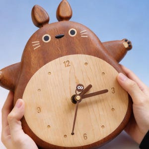 May include: A wooden clock shaped like a cartoon character with a light brown face and dark brown body. The clock face is light brown with dark brown hands and hour markers. The clock is approximately 8 inches tall.