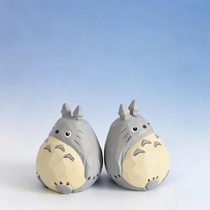 May include: Two gray and cream-colored Totoro figurines. Each figurine has a round body, pointed ears, and a cream-colored belly. The figurines are set against a gradient blue background.