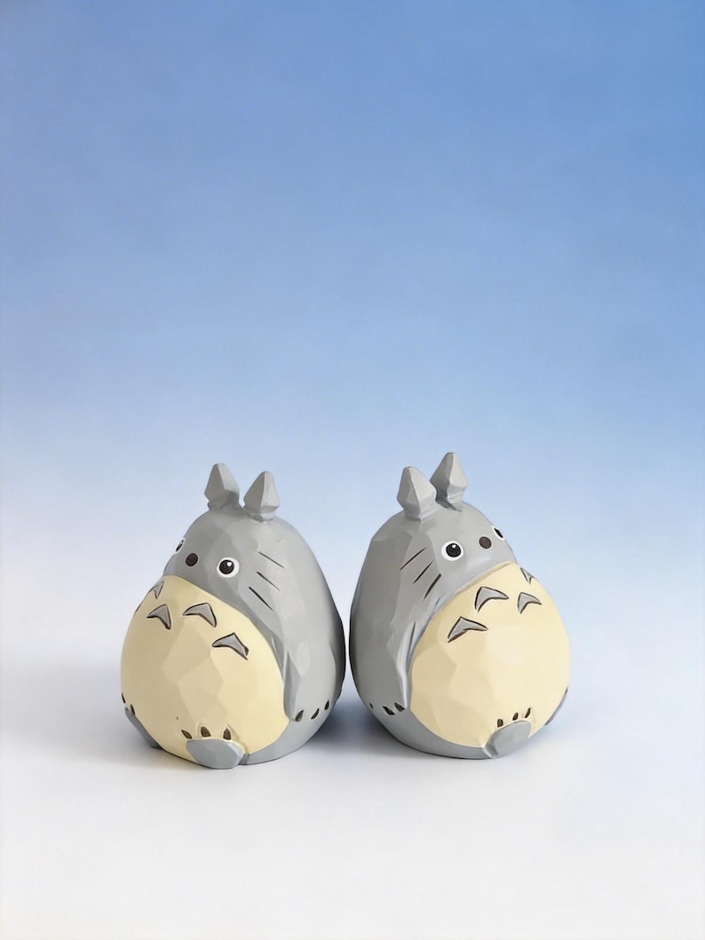 May include: Two gray and cream-colored Totoro figurines. Each figurine has a round body, pointed ears, and a cream-colored belly. The figurines are set against a gradient blue background.