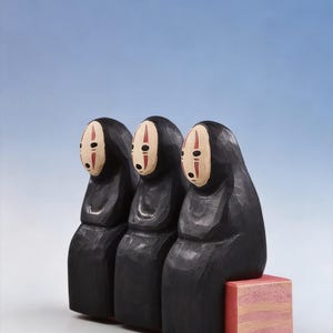 May include: Three black wooden figures with white and red facial details, resembling characters from a Japanese animated film. They sit on a small, pink, rectangular block against a gradient blue background. The figures have a hooded appearance.