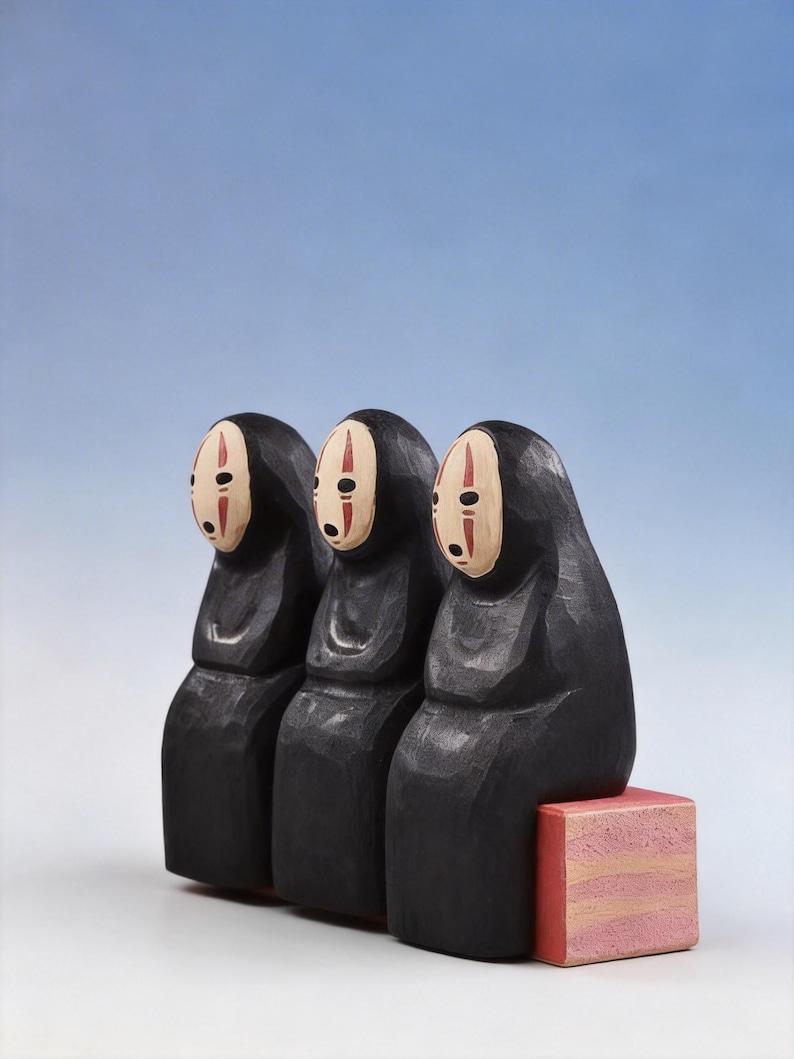 May include: Three black wooden figures with white and red facial details, resembling characters from a Japanese animated film. They sit on a small, pink, rectangular block against a gradient blue background. The figures have a hooded appearance.