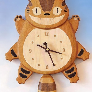 May include: A wooden wall clock shaped like a grinning cat character with a light-colored circular face, dark brown stripes, and a swinging pendulum tail. The clock features black hands, numerals at 3, 6, 9, and 12, and small dots for other hours.