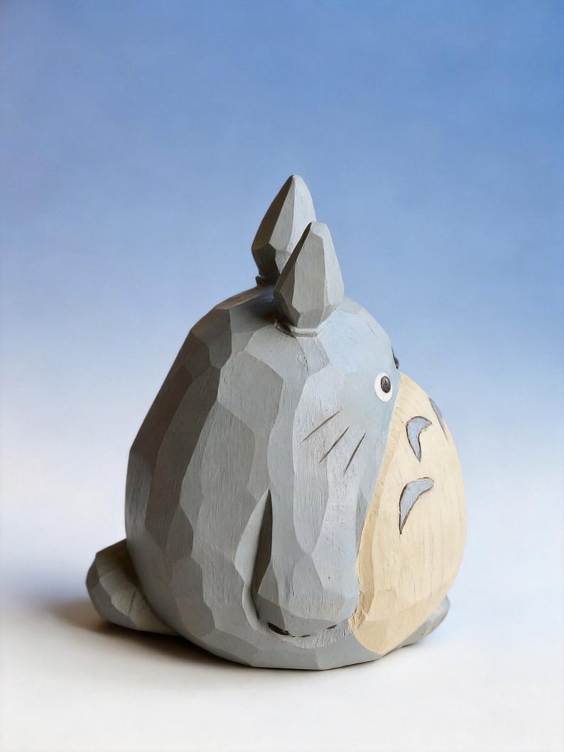 May include: A gray wooden sculpture of a cartoon character with large ears and a round body. The figure has a light beige belly and a small, black eye. The sculpture is carved with visible wood grain.