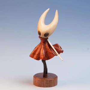 以下が含まれることがあります： 明るい色の頭と湾曲した角、暗い目、赤いスカートを持つキャラクターの木製フィギュア。フィギュアは細い、明るい色の木の杖を持っています。フィギュアは丸い、濃い色の木の台座に取り付けられています。