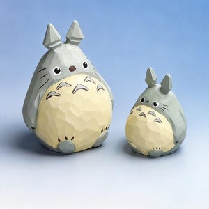 May include: Two wooden Totoro figurines. The larger one is gray with a cream-colored belly and black markings. The smaller one has the same color scheme. Both have pointed ears and are set against a blue gradient background.