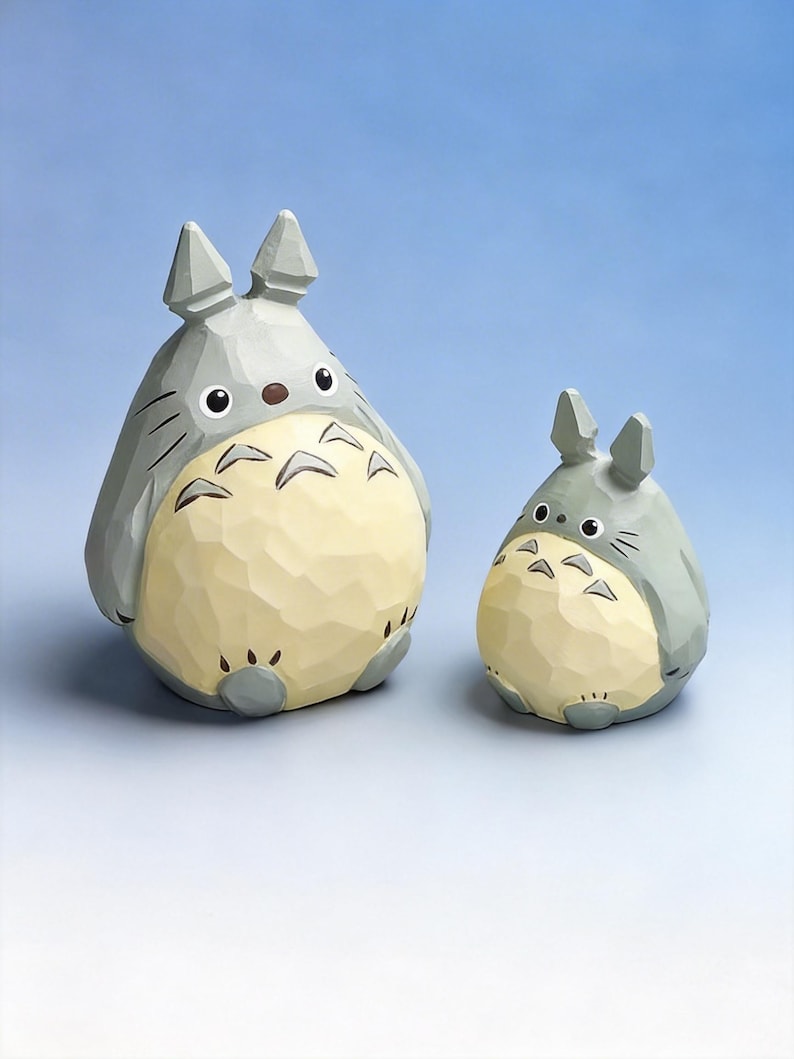 May include: Two wooden Totoro figurines. The larger one is gray with a cream-colored belly and black markings. The smaller one has the same color scheme. Both have pointed ears and are set against a blue gradient background.