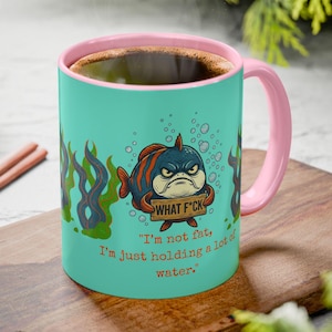 May include: A teal ceramic mug with a pink interior and handle. The mug features a cartoon fish with an angry expression holding a sign that says "WHAT F*CK". The text on the mug reads, "I'm not fat, I'm just holding a lot of water."