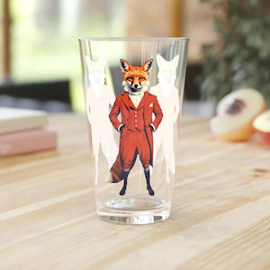 Puede incluir: Un vaso transparente con un zorro vestido con un traje rojo, flanqueado por siluetas blancas de zorros. El zorro está representado con las manos en los bolsillos. El vaso está sobre una mesa de madera.
