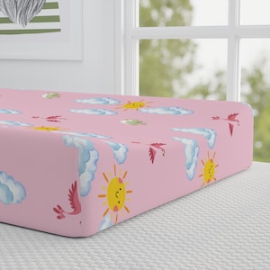 Könnte beinhalten: Rosa Spannbettlaken mit einem sich wiederholenden Muster aus lächelnden Sonnen, flauschigen blauen Wolken und rosa Vögeln. Das Laken ist für ein Babybett konzipiert und aus weichem Stoff gefertigt.