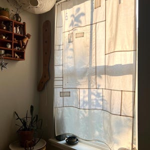May include: A sheer off-white window curtain featuring a grid of stitched rectangular panels and linear seams. The fabric is translucent, allowing sunlight to filter through and cast shadows of outdoor foliage onto the surface of the curtain.