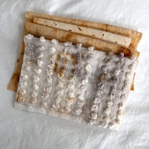 May include: A stack of handmade fabric squares in shades of beige and white. The top layer features small, raised, knotted details. The layers below have a more open weave with decorative cut-out holes. The fabrics are arranged on a white surface.