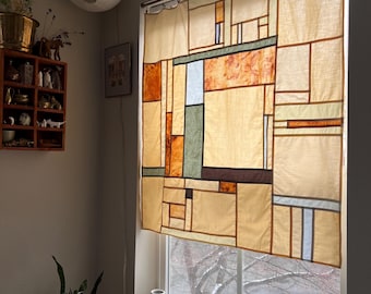 Puede incluir: Una persiana beige con un diseño geométrico de rectángulos y cuadrados en tonos marrón, naranja, verde y azul. La persiana cuelga frente a una ventana, con vistas a un árbol exterior. La habitación tiene una combinación de colores neutros.