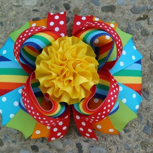 May include: A vibrant, handmade hair bow featuring a bright yellow center surrounded by layers of colorful ribbons. The ribbons include rainbow stripes, polka dots, and solid colors like red, orange, green, and blue. This accessory is perfect for adding a pop of color.