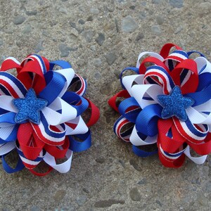 May include: Two red, white, and blue ribbon hair bows with a blue glitter star in the center. The bows are perfect for a patriotic celebration.