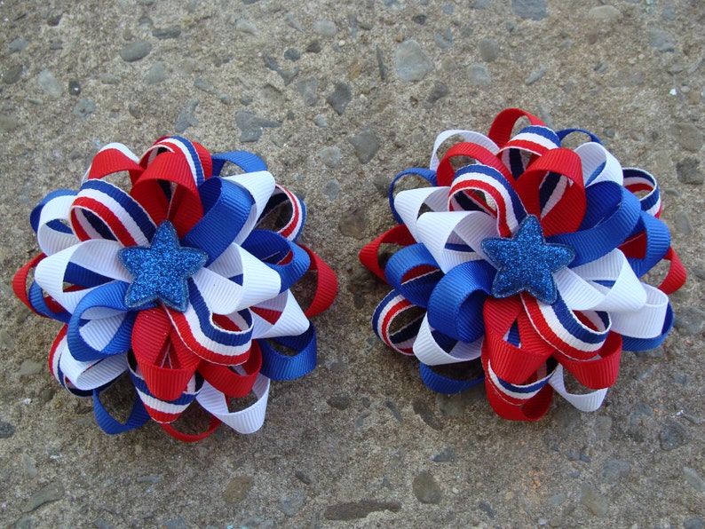 May include: Two red, white, and blue ribbon hair bows with a blue glitter star in the center. The bows are perfect for a patriotic celebration.