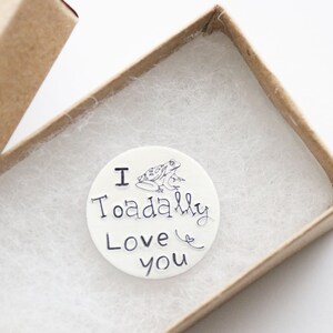 May include: A white round ceramic ornament with a black and white illustration of a frog and the text "I Toaddally Love You". The ornament is in a small brown box with white tissue paper.