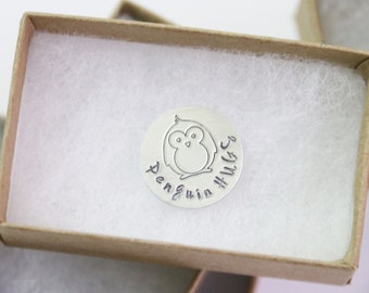 May include: A silver charm with a penguin design and the text "Penguin Hugs" engraved on it. The charm is in a small brown box with white padding.