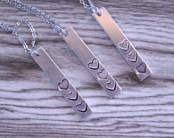 May include: Three silver necklaces with bar pendants featuring different numbers of engraved hearts. The pendants are on a wood background.
