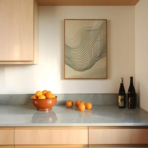 Könnte beinhalten: Ein gerahmtes, rechteckiges Wandbild mit dunkelgrünen, wellenförmigen Linienmustern auf beigem Hintergrund, das über einer Küchentheke mit einer Terrakottaschale voller Orangen und zwei dunklen Glasflaschen auf der rechten Seite hängt.