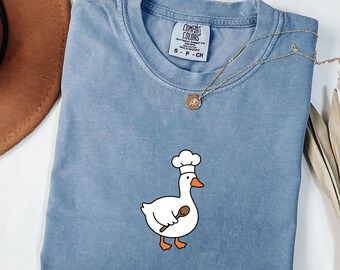 May include: A blue Comfort Colors t-shirt featuring a cartoon goose wearing a chef's hat and holding a wooden spoon. The shirt has a crew neck and short sleeves. A gold necklace with a pendant is draped across the shirt.
