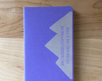 May include: A small, lavender notebook with the text "AIN'T NOBODY STOPPING ME I'M WALKING LIKE A TITAN" printed in white. The notebook has a geometric design on the upper right corner and is lying on a wooden surface.